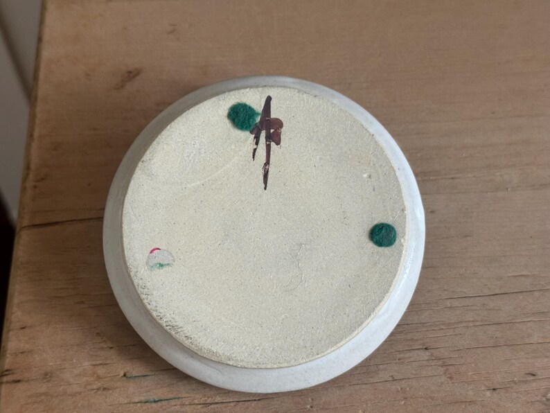 May include: A small, off-white ceramic dish with a slightly raised edge. The underside features a speckled texture and is decorated with a dark brown and green abstract design, along with two green and one pink dot.