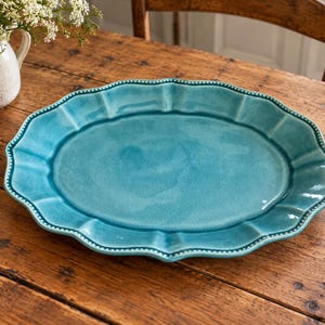 Könnte beinhalten: Eine ovale Servierplatte aus Keramik mit einer leuchtend türkisblauen Glasur. Die Platte hat einen gewellten Rand mit einem feinen Perlenmuster entlang der Kante, das eine strukturierte Umrandung um die glatte, glänzende Oberfläche bildet.