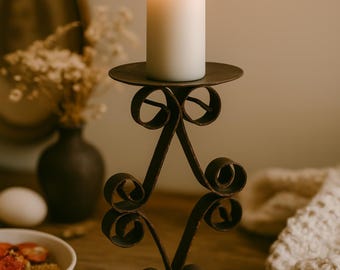 May include: A burning white candle on a decorative wrought-iron candle holder. The candle holder has a circular base and a scrolled design. The candle holder is made of dark metal and is on a wooden surface.