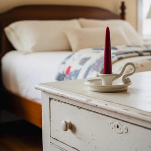 May include: A red taper candle in a white ceramic candlestick holder sits on a distressed white nightstand. The bedroom setting includes a bed with white linens and a patterned quilt. A lamp is visible in the background.