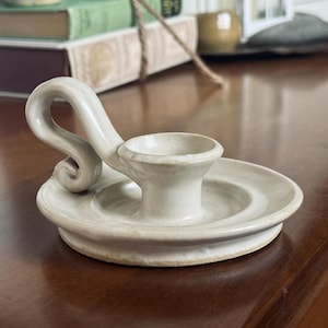 May include: Off-white ceramic candle holder with a circular base and a curved handle. The candle holder has a small cup to hold a candle. The handle has a decorative swirl. The candle holder is on a dark brown surface.