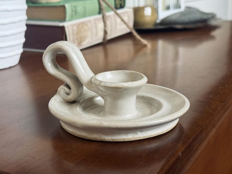 May include: Off-white ceramic candle holder with a circular base and a curved handle. The candle holder has a small cup to hold a candle. The handle has a decorative swirl. The candle holder is on a dark brown surface.