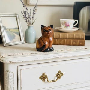 Op de afbeelding: Een houten kattenbeeldje met zwarte accenten staat op een witte dressoir. Een ingelijste foto, een vaasje met lavendel en een kop en schotel zijn ook aanwezig. De dressoir heeft sierlijke messing hardware.