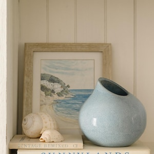 Peut inclure: Un vase en céramique bleu clair avec une ouverture inclinée unique est posé sur une étagère avec deux coquillages et deux livres. Les livres indiquent "VINTAGE BEMIXED CF" et "SUNNYLANDS". Une peinture encadrée d'une scène côtière est en arrière-plan.