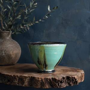 May include: A ceramic bowl with a flared rim and a pedestal base, featuring a vibrant mint green glaze with vertical streaks of dark brown and yellow. The bowl has a smooth finish and a dark, contrasting rim, showcasing a hand-thrown appearance.