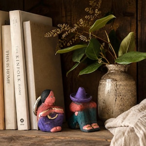 May include: Two small clay figurines sit on a shelf. One figure has black hair, a red top, and a purple skirt with gold accents. The other figure wears a purple sombrero, a red shirt, and teal trousers with gold details. A speckled ceramic vase holds green leaves.