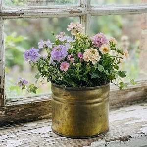 Peut inclure: Un récipient en métal couleur laiton rempli d'une variété de fleurs colorées, dont des fleurs violettes, roses et crème. Le récipient est posé sur un rebord de fenêtre patiné peint en blanc, avec un arrière-plan vert flou visible à travers la fenêtre.
