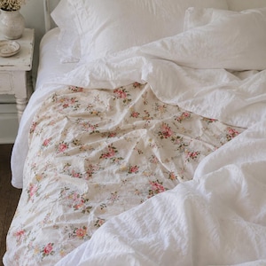 May include: A cream-coloured throw blanket featuring a delicate floral pattern with small pink, yellow, and red roses and green leaves. The blanket is draped over a bed with white bedding, including a white duvet cover and white pillows.