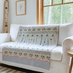 Puede incluir: Una manta beige y azul estampada sobre un sofá blanco. La manta presenta un diseño geométrico con formas azules y negras y zigzags rojos. El sofá está sobre una alfombra a rayas, cerca de una ventana con vista al bosque.