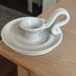 May include: Off-white ceramic candle holder with a circular base and a curved handle. The candle holder has a small cup to hold a candle and is sitting on a wooden surface.