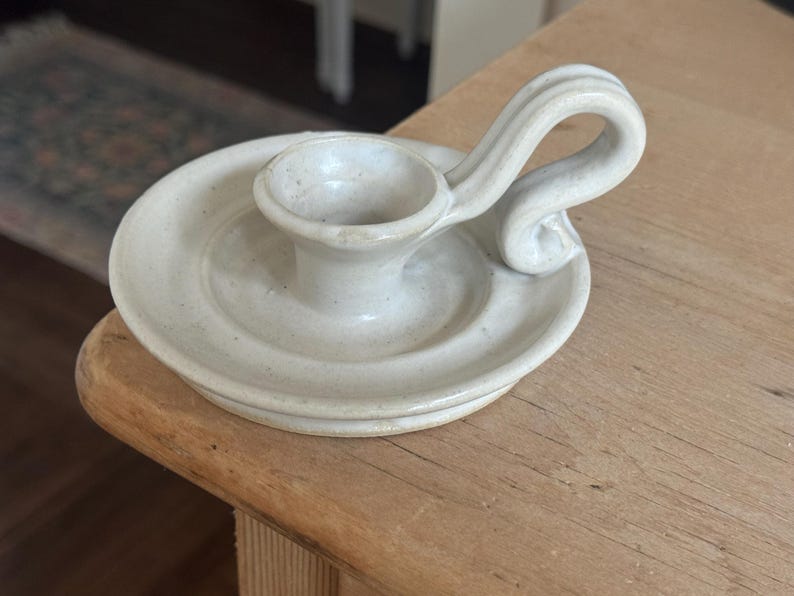 May include: Off-white ceramic candle holder with a circular base and a curved handle. The candle holder has a small cup to hold a candle and is sitting on a wooden surface.