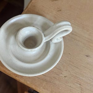 May include: A handmade, off-white ceramic candle holder. The candle holder has a circular base with a raised center for the candle and a curved handle. The piece is placed on a wooden surface.