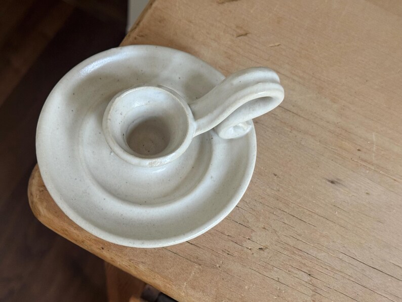 May include: A handmade, off-white ceramic candle holder. The candle holder has a circular base with a raised center for the candle and a curved handle. The piece is placed on a wooden surface.