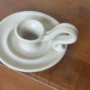 May include: A handmade, off-white ceramic candle holder. The candle holder has a circular base with a raised center for the candle and a curved handle. The piece has a rustic, speckled appearance.