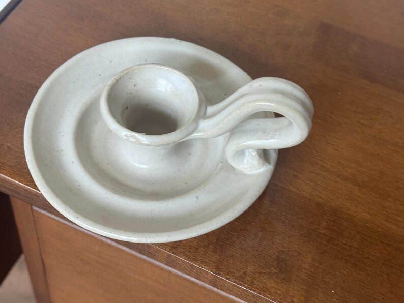 May include: A handmade, off-white ceramic candle holder. The candle holder has a circular base with a raised center for the candle and a curved handle. The piece has a rustic, speckled appearance.
