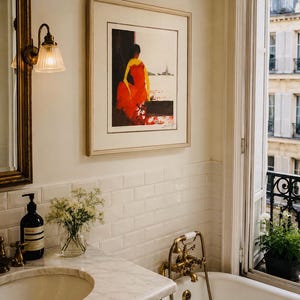 May include: A framed art print featuring a stylised illustration of a figure in a vibrant red dress against a dark background, displayed on a wall above a white marble vanity with a sink, a soap dispenser, and a small glass vase of flowers.