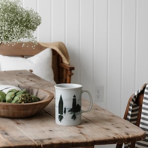 Puede incluir: Una taza de cerámica blanca con una escena costera en blanco y negro que presenta un faro y árboles. La taza está sobre una mesa de madera rústica, junto a un cuenco de madera con alcachofas y una toalla a rayas.