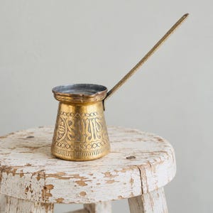 May include: A brass coffee pot with a long, slender handle and a wide, flat base. The exterior features intricate, hand-engraved patterns including geometric borders and stylized floral motifs. The interior rim shows a metallic, silver-toned finish.