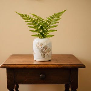 May include: A white ceramic vase with a botanical pattern, filled with green fern leaves. The vase is placed on a dark wooden table with a drawer. The room has a warm, neutral colour scheme.