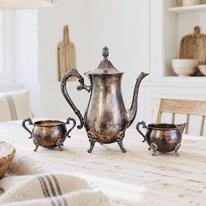 May include: A silver-plated tea set comprising a tall teapot with a lid, a sugar bowl, and a creamer. Each piece features a tarnished metallic finish, ornate curved handles, and decorative feet. The teapot has a pointed finial on the lid.
