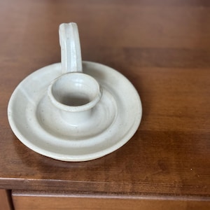 May include: Off-white ceramic candle holder with a circular base and a small, raised cup to hold a candle. It has a curved handle on the side. The candle holder is sitting on a wooden surface.