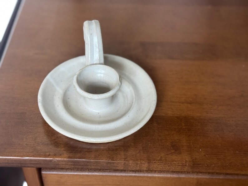 May include: Off-white ceramic candle holder with a circular base and a small, raised cup to hold a candle. It has a curved handle on the side. The candle holder is sitting on a wooden surface.