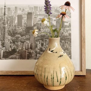 May include: A beige ceramic vase with a rounded form, adorned with green vertical strokes and small black bird motifs. The vase contains a sprig of purple flowers, white daisies, and pink coneflowers. A framed monochrome cityscape is in the background.