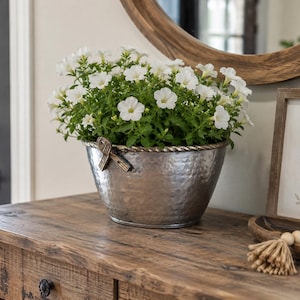 Op de afbeelding: Een zilverkleurige, gehamerde metalen plantenbak met een touwachtige rand en decoratieve zijaccenten, gevuld met een weelderig arrangement van witte petunia's. De plantenbak heeft een getextureerd oppervlak en een taps toelopende vorm.