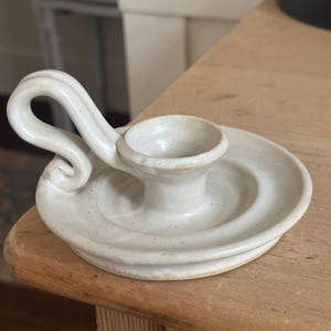May include: Off-white ceramic candle holder with a circular base and a curved handle. The candle holder has a small, central cup for the candle and is placed on a wooden surface. The design is simple and rustic.