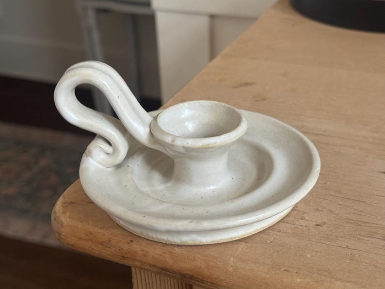 May include: Off-white ceramic candle holder with a circular base and a curved handle. The candle holder has a small, central cup for the candle and is placed on a wooden surface. The design is simple and rustic.