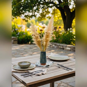 May include: An outdoor dining table set with a vase of dried flowers, plates, bowls, and cutlery. The vase is blue and brown. The plates and bowls are green. The table is set on a stone patio with a blurred background of trees and flowers.
