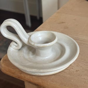 May include: Off-white ceramic candle holder with a circular base and a central cup for the candle. It features a curved handle. The piece is handmade and has a rustic, speckled appearance. A decorative item for home decor.