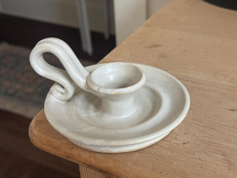 May include: Off-white ceramic candle holder with a circular base and a central cup for the candle. It features a curved handle. The piece is handmade and has a rustic, speckled appearance. A decorative item for home decor.