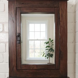 May include: A dark brown wooden medicine cabinet with a rectangular mirror. The cabinet has a rustic, distressed finish and a small shelf at the bottom. A small plant is visible inside the mirror's reflection, in front of a window.