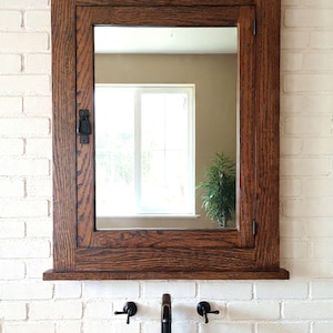 May include: A rectangular wooden framed mirror with a cabinet door, mounted on a white brick wall. The mirror is above a white ceramic sink with black fixtures. The wood has a rich, dark brown finish.