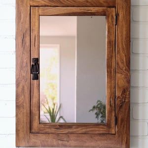 Puede incluir: Un espejo rectangular con marco de madera y puerta. La madera tiene un tinte marrón cálido y veta visible. La puerta tiene un pestillo de metal negro. El espejo está montado en una pared de ladrillo blanco.
