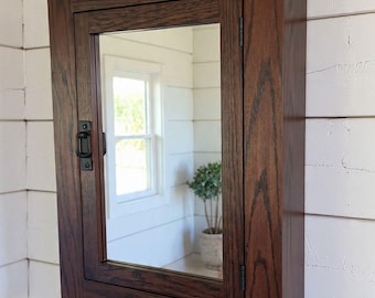May include: A dark brown wooden medicine cabinet with a mirrored door. The cabinet is rectangular with a small shelf at the bottom and is mounted on a white wall. The door has a black latch and hinges. The wood grain is visible.