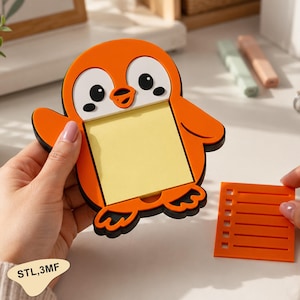 以下が含まれることがあります： 黒い輪郭を持つオレンジ色のペンギン型付箋ホルダーで、中央に正方形の黄色いメモ帳が付いています。その横には、水平線と正方形の切り抜きが縦一列に並んだ、小さなオレンジ色の長方形のプラスチック製ステンシルが置かれています。
