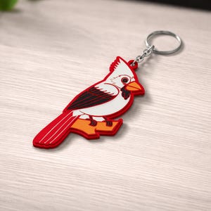May include: A red, white, and black cardinal bird keychain. The keychain features a detailed design of the bird perched on a branch, with a silver metal keyring attached. The bird's feathers are red, with black accents and a white body.