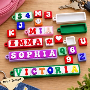 Può includere: Una collezione di ciondoli colorati in silicone a forma di lettere e numeri per portachiavi, con nomi come EMMA, SOPHIA e VICTORIA, oltre a singole lettere, numeri, un'icona di baseball, un cuore e un fiore.