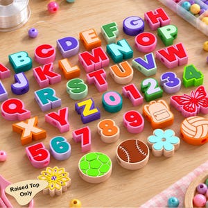 May include: A collection of colorful silicone beads featuring letters of the alphabet, numbers zero through nine, and shapes including a butterfly, backpack, flower, soccer ball, baseball, and volleyball, with a label stating Raised Top Only.