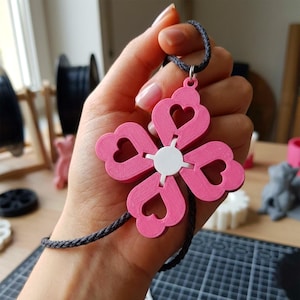 以下が含まれることがあります： ハート型の切り抜きと白い中心部を備えたピンク色の花のペンダントネックレス。ペンダントは濃い灰色のコードから吊り下げられています。ネックレスは手で持たれています。