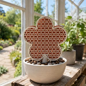 May include: A decorative flower-shaped garden stake with a white outline and a patterned reddish-brown center. The stake is placed in a white ceramic pot filled with pebbles. The pot sits on a wooden surface near a window.