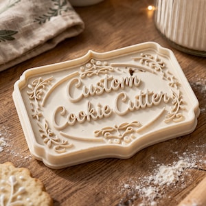 May include: A beige rectangular cookie cutter with the words "Custom Cookie Cutter" in a cursive font. The cutter has a decorative floral border and is on a wooden surface with flour.