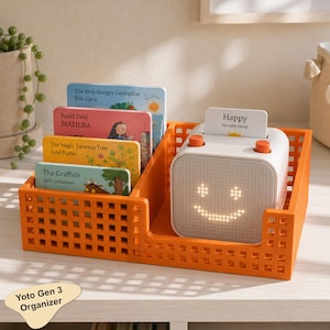 Puede incluir: Una cesta de almacenamiento de plástico naranja con patrón de rejilla que contiene un reproductor de audio Yoto blanco con una cara sonriente brillante y una ranura para tarjetas. Cuatro libros infantiles están en el compartimento adyacente, incluyendo The Very Hungry Caterpillar, Matilda, The Magic Faraway Tree y The Gruffalo.
