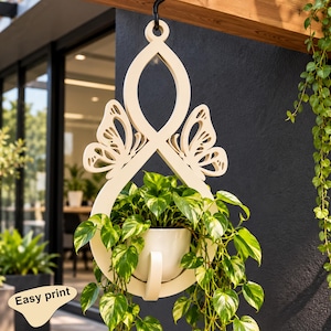 May include: A cream-coloured hanging plant holder featuring a central loop design with two symmetrical butterfly cutouts on the sides. The structure supports a small, plain white ceramic pot containing a trailing green and yellow pothos plant.