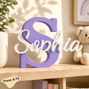 Può includere: Un'insegna decorativa in legno caratterizzata da una grande lettera S maiuscola viola con il nome Sophia scritto in lettere minuscole bianche in corsivo sovrapposte al centro della lettera S, creando un aspetto dimensionale e stratificato.