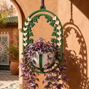 May include: A dark green oval hanging plant holder featuring a decorative frame with small cream-colored heart accents and leaf-shaped cutouts. A white bowl-shaped planter sits inside the frame, containing a trailing plant with purple and green striped leaves.