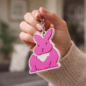 Puede incluir: Un llavero de conejo rosa con contorno blanco y anilla plateada. El conejo es un diseño estilizado con una estética sencilla y moderna. El llavero se sostiene sobre un fondo borroso.