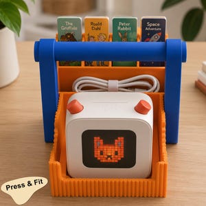 Puede incluir: Un reproductor de audio digital cuadrado de color blanco con una cara de gato naranja pixelada en la pantalla y dos perillas naranjas en la parte superior, dentro de un organizador de plástico naranja con asa azul. Incluye cuatro tarjetas y un cable blanco.